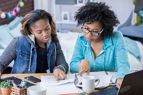 Two teen-girls-writing-on-a-resume-together with papers and a laptop in front of them.